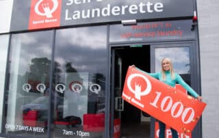 A blonde woman stands holding a sign that reads "Self Service Launderette," promoting laundry services in a public space
